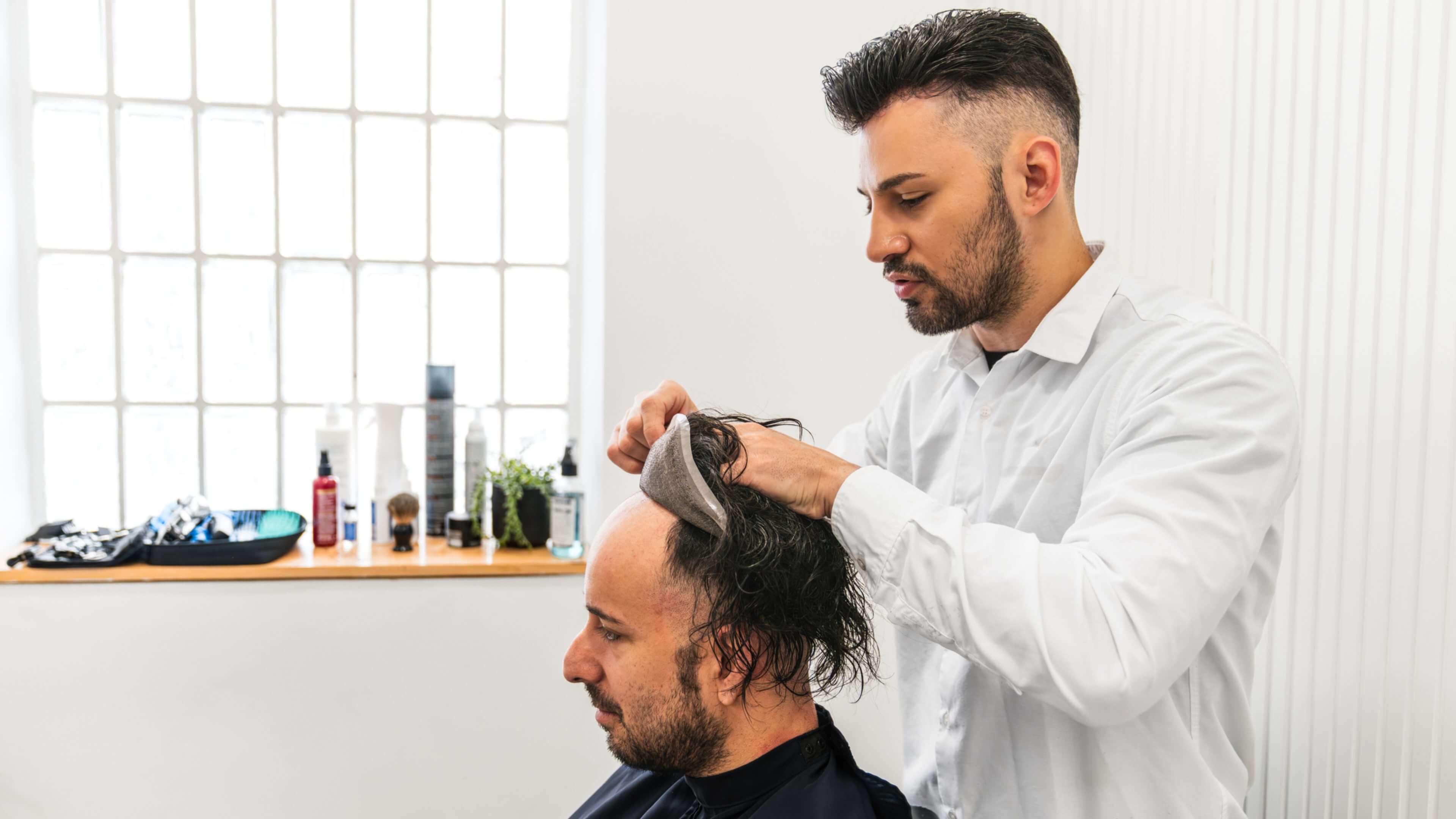 Man fitting hairpiece on another man's head.