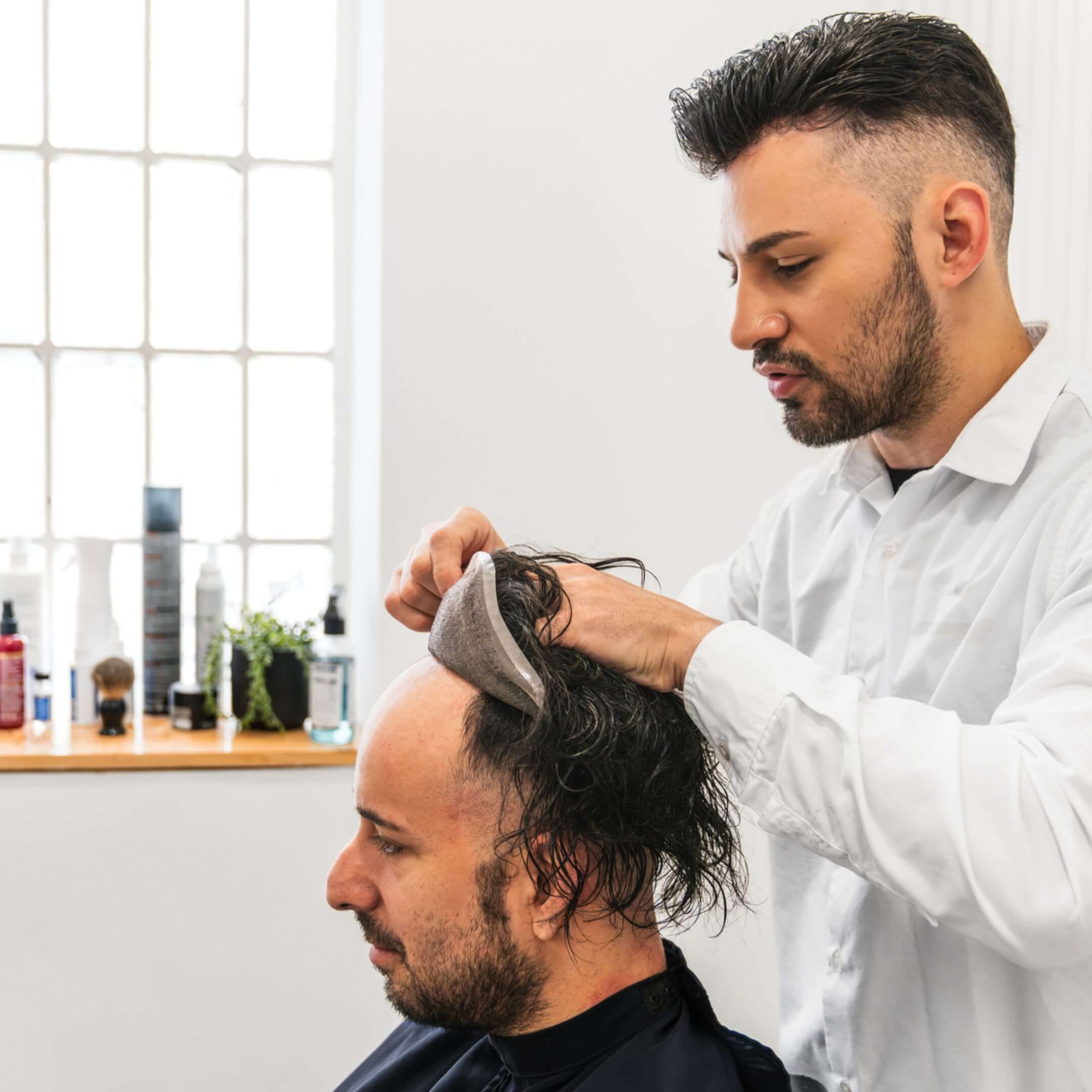 Man fitting hairpiece on another man's head.